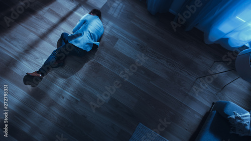 Poor Depressed Drunk Young Man is Sleaping on the Floor Near Sofa in an Apartment with Wooden Flooring. Dramatic Top View Camera Shot. photo