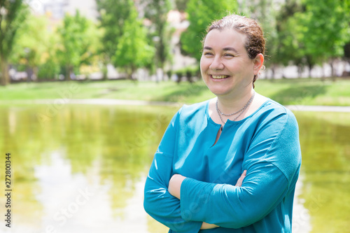 Cheerful woman enjoying outdoor walk and laughing. Happy mid adult Caucasian lady with arms crossed standing in park near pond, looking away and smiling. Leisure time outdoors concept
