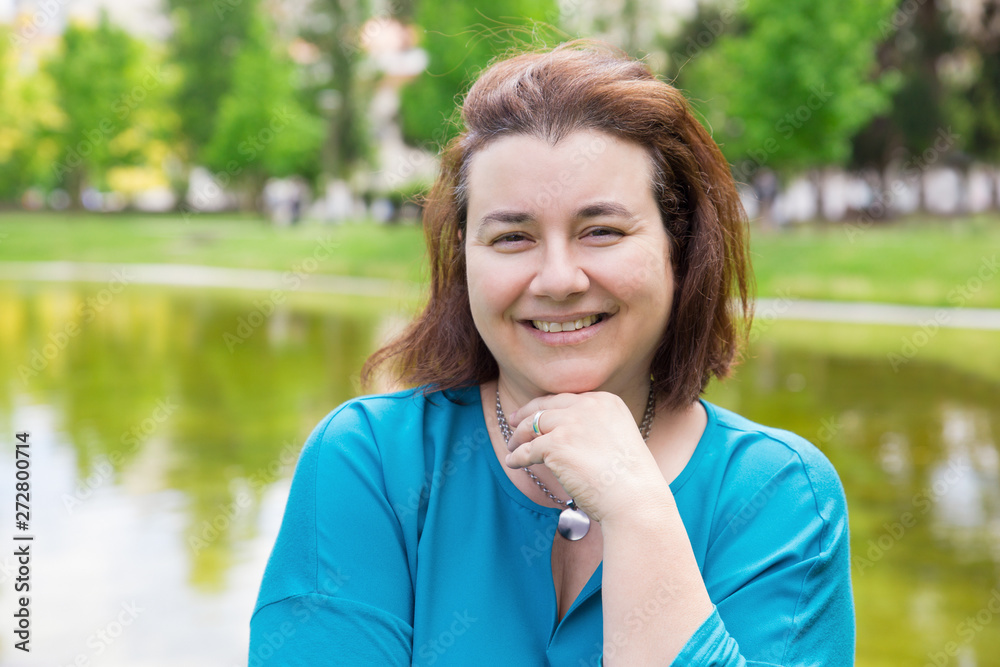 Happy cheerful woman enjoying weekend and leisure time outdoors. Middle aged Caucasian lady in casual standing in park near pond and smiling at camera. Outdoor female portrait concept Happy cheerful woman enjoying weekend and leisure time outdoors. Middle aged Caucasian lady in casual standing in park near pond and smiling at camera. Outdoor female portrait concept