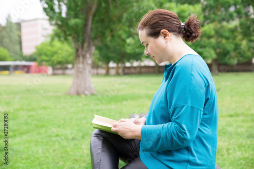 Relaxed female book lover enjoying novel outdoors. Side view of calm Caucasian lady sitting in park and reading. Hobby concept