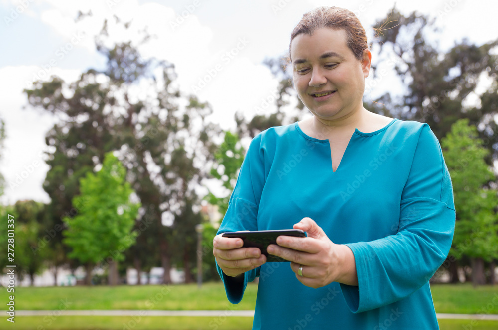 Smiling pensive woman watching media content on phone outdoors. Middle aged Caucasian lady using smartphone while walking in park. Wireless connection concept Smiling pensive woman watching media content on phone outdoors. Middle aged Caucasian lady using smartphone while walking in park. Wireless connection concept