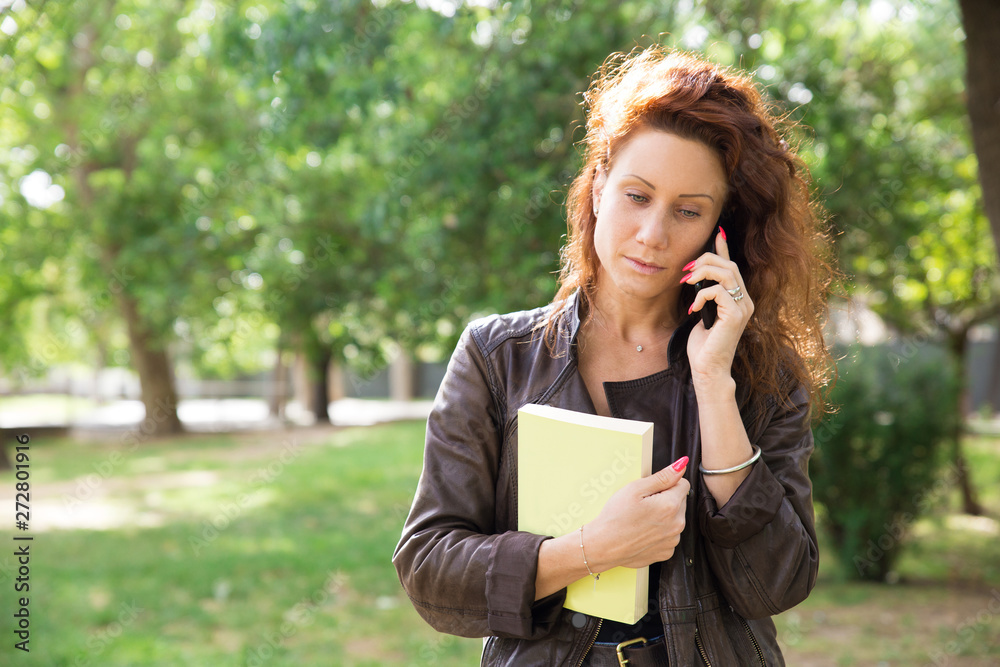 Serious young woman talking on phone. Pensive pretty brunette walking on campus with book and making phone call. Communication concept Serious young woman talking on phone. Pensive pretty brunette walking on campus with book and making phone call. Communication concept
