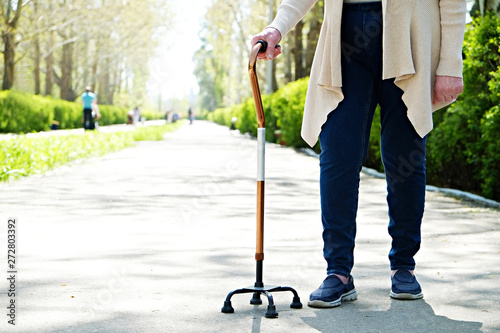 Senior disabled caucasian woman hands on cane outside nursing home park. Close up of elderly lady holding a walking stick outdoors of healthcare facility on the sunny day.