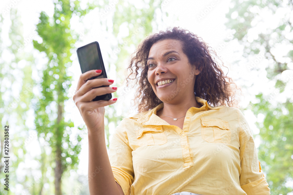 Surprised woman browsing on smartphone in city park. Young lady looking at gadget screen with blurred green trees in background. Surprise concept. Front view.