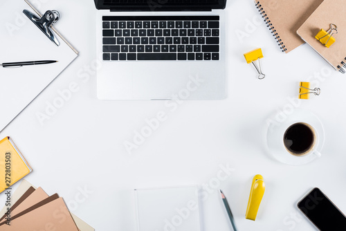 top view of laptop, clipboard, craft paper, notebooks, smartphone with blank screen, binder clips, book and cup of coffee on white
