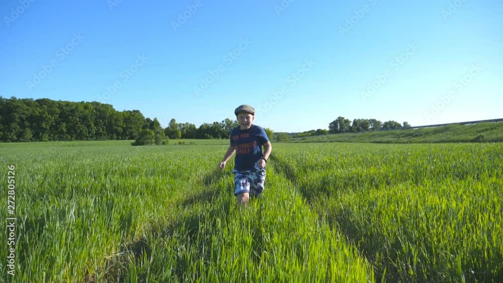 Portrait of happy boy is running through the field with green wheat at a sunny hot day. Smiling child in hat jogging at the meadow in summer and having fun. Concept of childhood and rest. Slow motion Portrait of happy boy is running through the field with green wheat at a sunny hot day. Smiling child in hat jogging at the meadow in summer and having fun. Concept of childhood and rest. Slow motion
