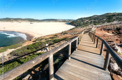Path on hills over ocean, made from wood, and empty windy sunny beach in Portugal town. Ocean waters and green hills over peaceful seaside at sunny day