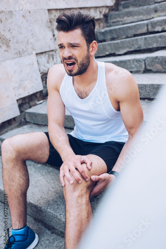 selective focus of injured sportsman screaming while sitting on stairs and suffering from pain