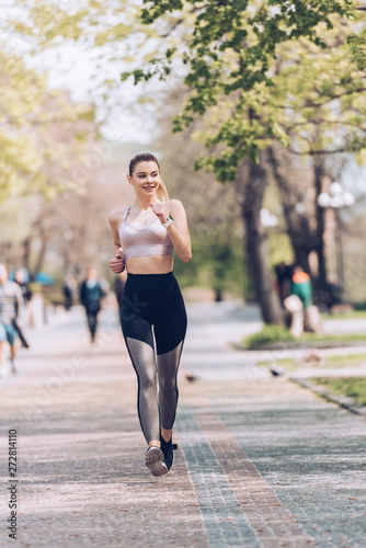 pretty sportsman smiling while jogging along wide walkway in park
