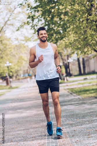 cheerful sportsman smiling while running along wide walkway in park