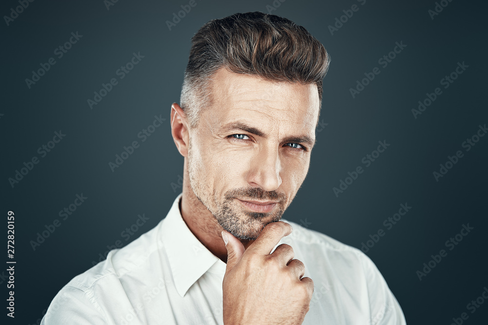 So charming! Handsome young man smiling and looking at camera while standing against grey background So charming! Handsome young man smiling and looking at camera while standing against grey background