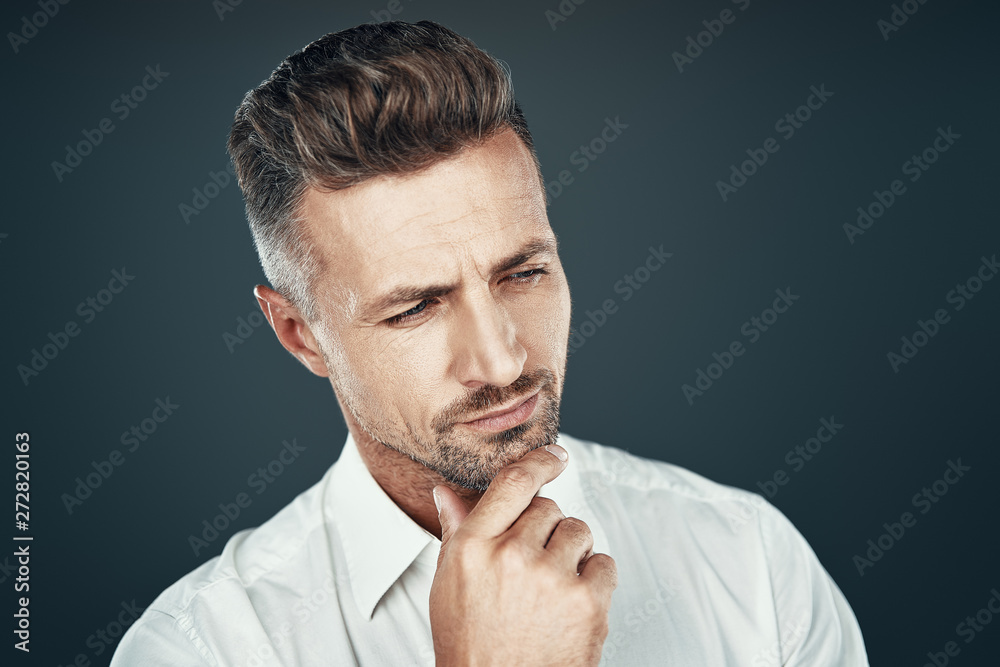 Male beauty. Handsome young man keeping hand on chin and looking away while standing against grey background