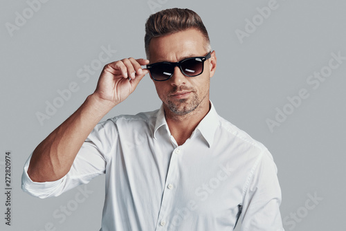 In his own style. Handsome young man looking at camera and adjusting sunglasses while standing against grey background