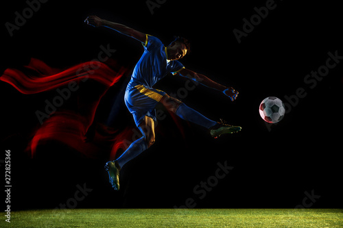 Young african-american male football or soccer player in sportwear and boots kicking ball for the goal in mixed light on dark background. Concept of healthy lifestyle  professional sport  hobby.