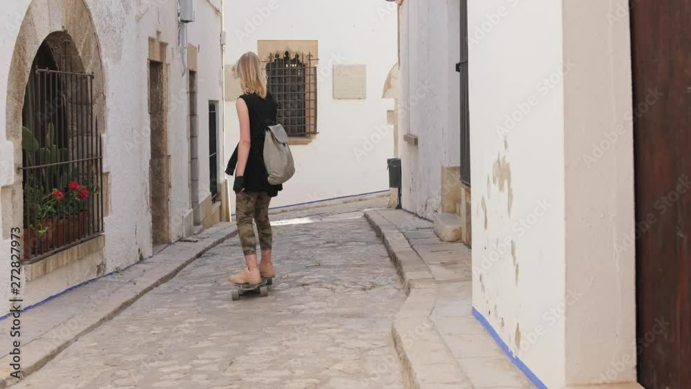 A beautiful blonde girl with backpack riding on skateboard through tight street A beautiful blonde girl with backpack riding on skateboard through tight street