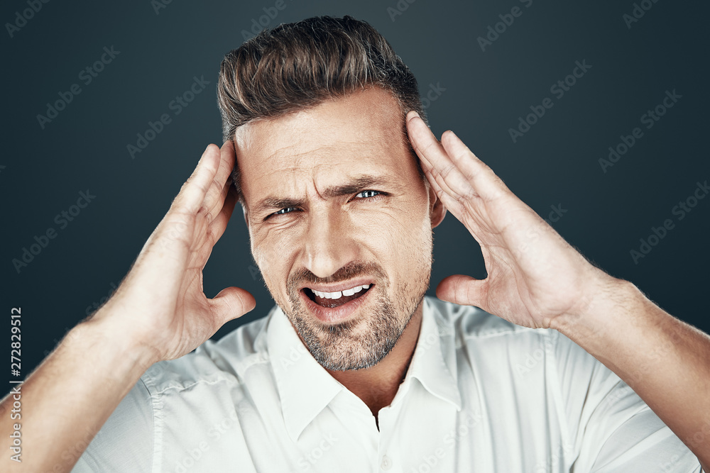 Headache. Frustrated young man keeping head in hands and making a face while standing against grey background Headache. Frustrated young man keeping head in hands and making a face while standing against grey background