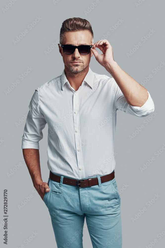 Modern style. Handsome young man looking at camera and adjusting sunglasses while standing against grey background Modern style. Handsome young man looking at camera and adjusting sunglasses while standing against grey background