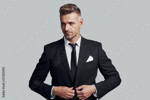 Young and successful. Handsome young man adjusting jacket and smiling while standing against grey background photo