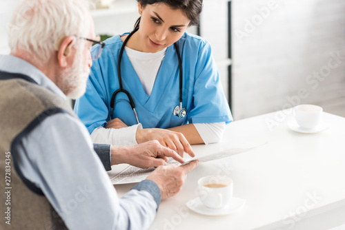 Cropped view of nurse looking at senior man with newspaper in hands