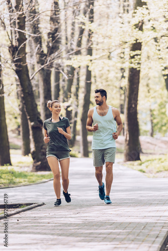 young man and woman in sportswear smiling while jogging in green park