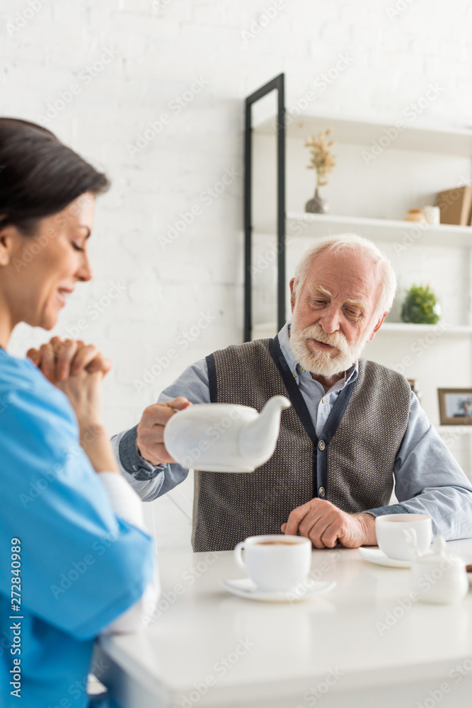Cheerful nurse sitting on kitchen at home, while senior man pouring tea in cup Cheerful nurse sitting on kitchen at home, while senior man pouring tea in cup