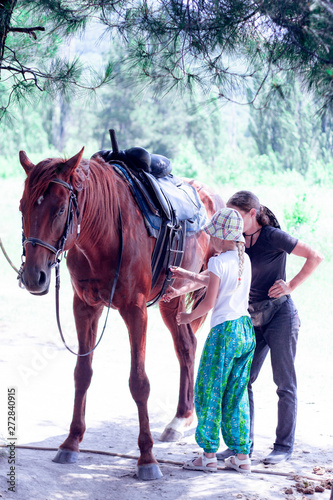 The instructor helps the girl to saddle a brown horse in the forest. Children's equestrian camp. Summer sports camp for children. A nice girl is learning to ride a horse.