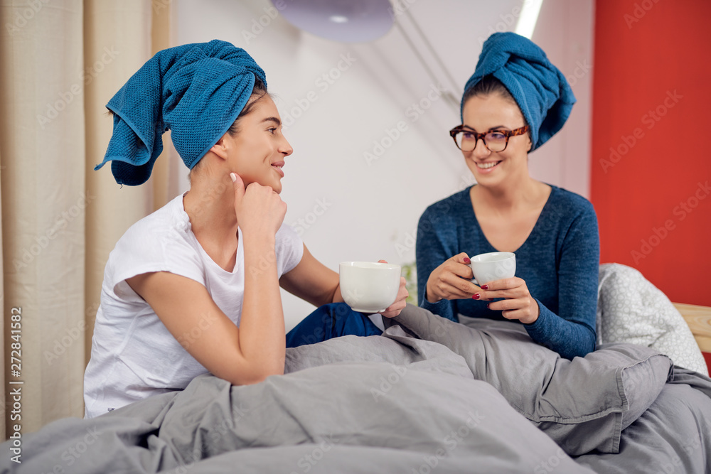 Teo beautiful Caucasian girlfriends with towels on heads sitting on bed in bedroom, drinking coffee and gossiping. Teo beautiful Caucasian girlfriends with towels on heads sitting on bed in bedroom, drinking coffee and gossiping.