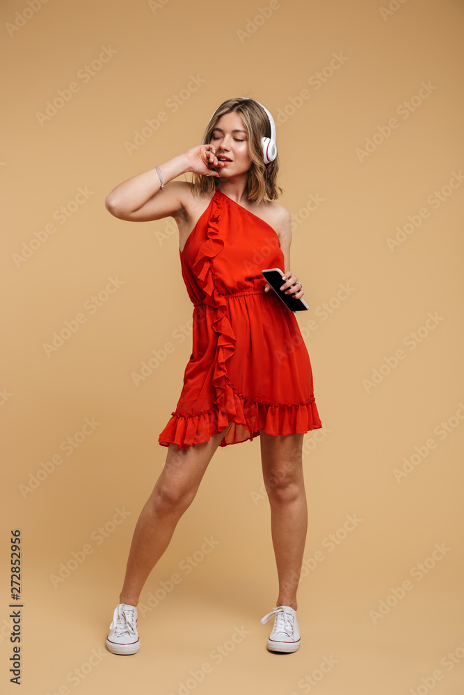 Full length photo of young woman wearing red dress holding cellphone and listening to music with earphones Full length photo of young woman wearing red dress holding cellphone and listening to music with earphones