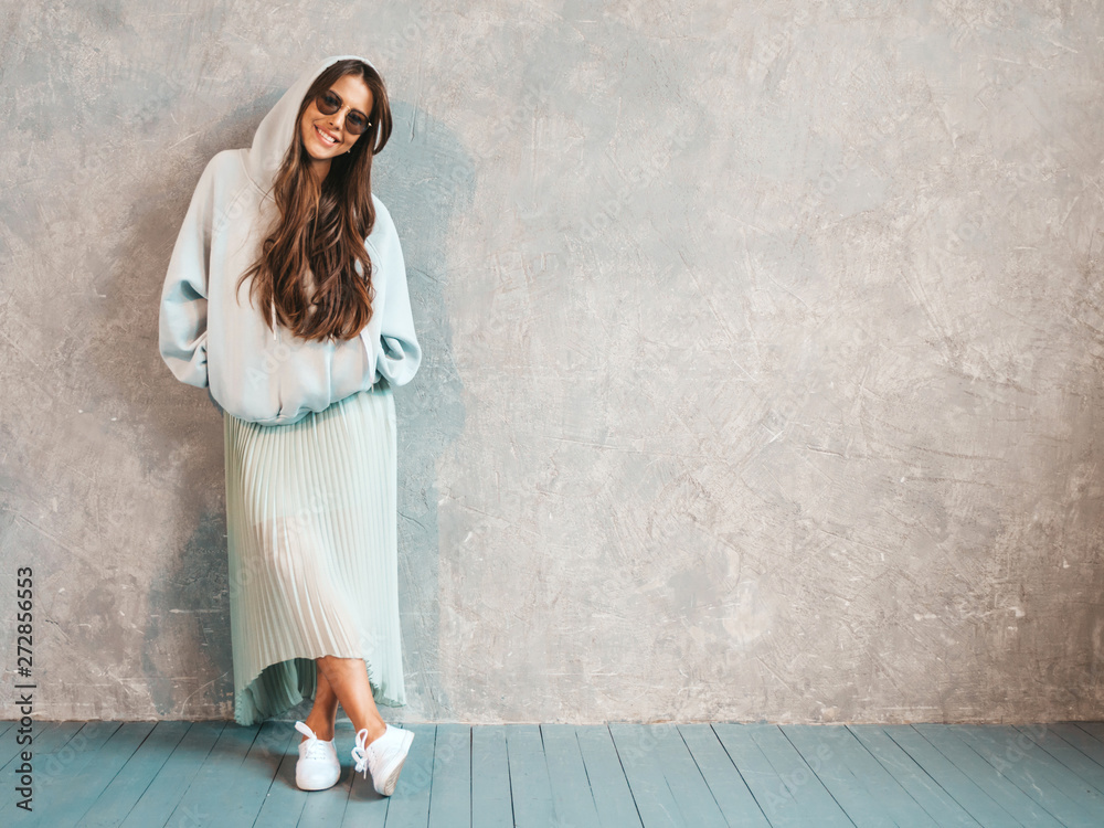 Young beautiful smiling woman looking at camera. Trendy girl in casual summer hoodie and skirt clothes. Funny and positive female posing near gray wall in studio. In sunglasses. Young beautiful smiling woman looking at camera. Trendy girl in casual summer hoodie and skirt clothes. Funny and positive female posing near gray wall in studio. In sunglasses.