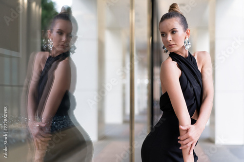 A beautiful European model is posing in a black dress near a shop window. One can see her reflection. Stylish atmosphere for advertising fashion trends. Average plan