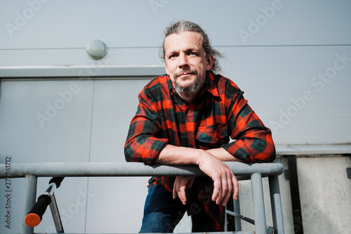 An elderly hipster man with long hair and a beard in a red plaid shirt leans on the railing against a gray building.