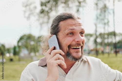 An elderly handsome man with long hair in the tail emotionally speaks on a cell phone in a park on a bench.