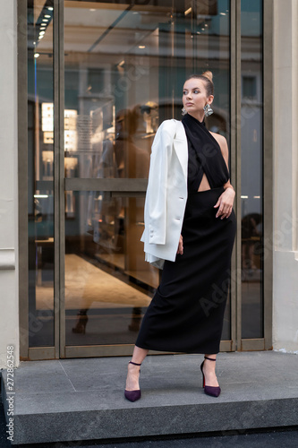 beautiful european model in black long dress and white jacket on her shoulders posing beside a boutique