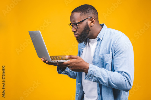 Young surprised african american man standing and using laptop computer isolated over yellow background.