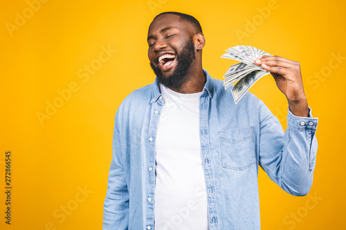 Winner! Young rich african american man in casual t-shirt holding money dollar bills with surprise isolated over yellow wall. photo