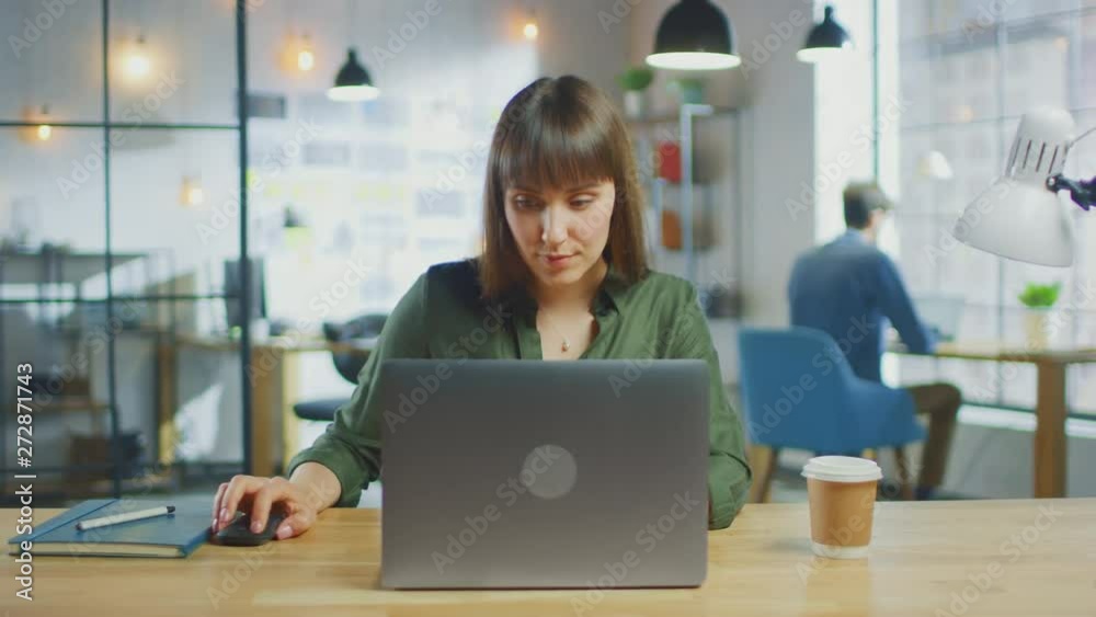 Young Beautiful Brunette Works on a Laptop Computer in Cool Creative Agency in a Loft Office. She has a Take-away Coffee and a Notebook on Her Table. Her Colleague Works in the Background. Young Beautiful Brunette Works on a Laptop Computer in Cool Creative Agency in a Loft Office. She has a Take-away Coffee and a Notebook on Her Table. Her Colleague Works in the Background.