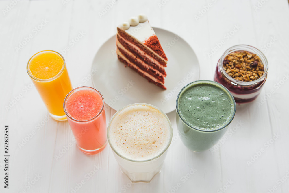 a red velvet cake on the background of glasses with various colored drinks (juice, coffee, smppthie, milkshake) standing on a white wooden table. A concept for a baker's dessert, a cafe, a restaurant a red velvet cake on the background of glasses with various colored drinks (juice, coffee, smppthie, milkshake) standing on a white wooden table. A concept for a baker's dessert, a cafe, a restaurant