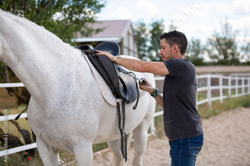 Side view of young guy putting black saddle on back of white horse while standing in paddock on ranch