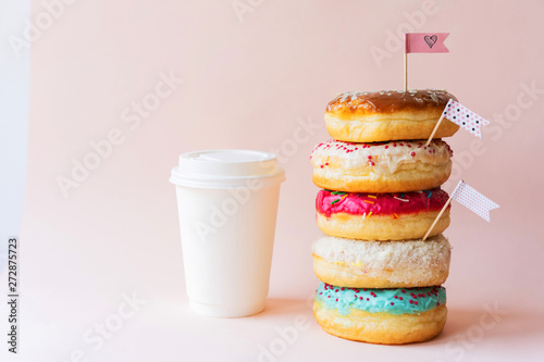 The colored glazed appetizing donuts are assembled in a tower and decorated with flag-shaped toppers, the paper cup is ideally white as a mock-up for text. Pink background with copy space.
