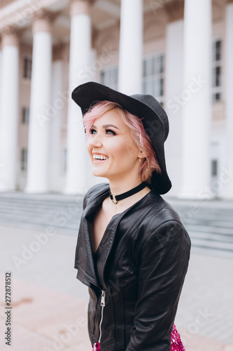 Young pretty positive sexy caucasian girl walking in the street in sexy dress and stylish black hat with pink hair smiling and having a nice day. Beauty,success, education concept