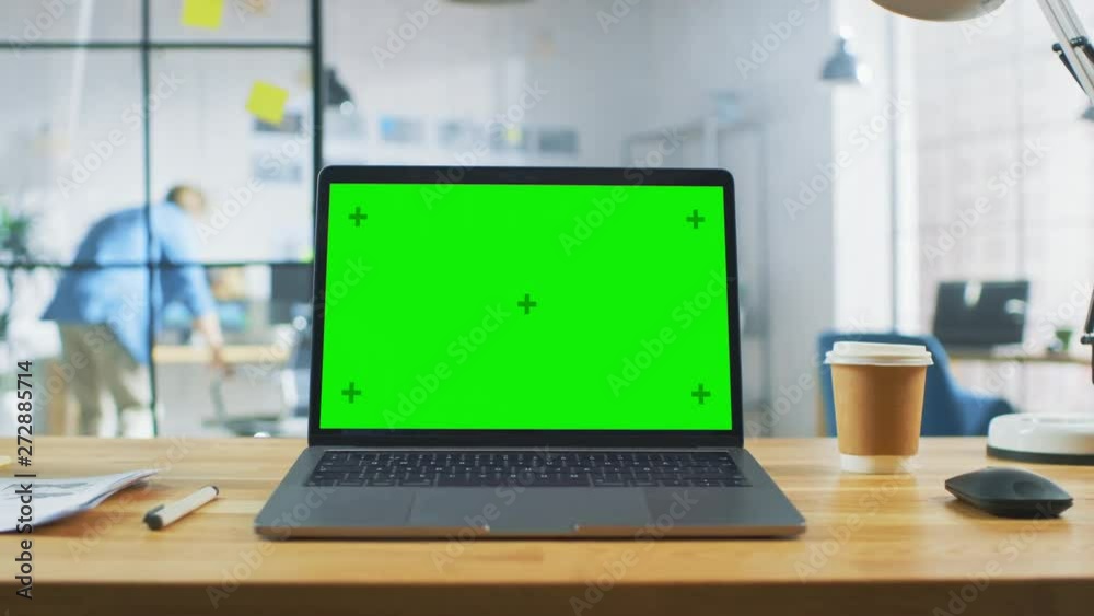Laptop on the Desk in the Office Shows Green Mock-up Screen. In the Background Bright Creative Open Space Loft Office HUB with Professional Working
