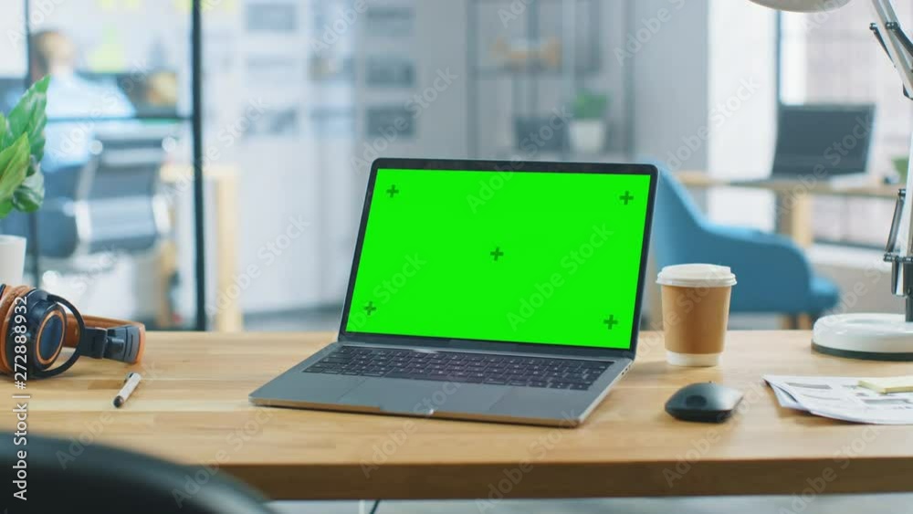 Laptop on the Desk in the Office Shows Green Mock-up Screen. In the Background Bright Modern Open Space Business Loft Office HUB with Professional Working