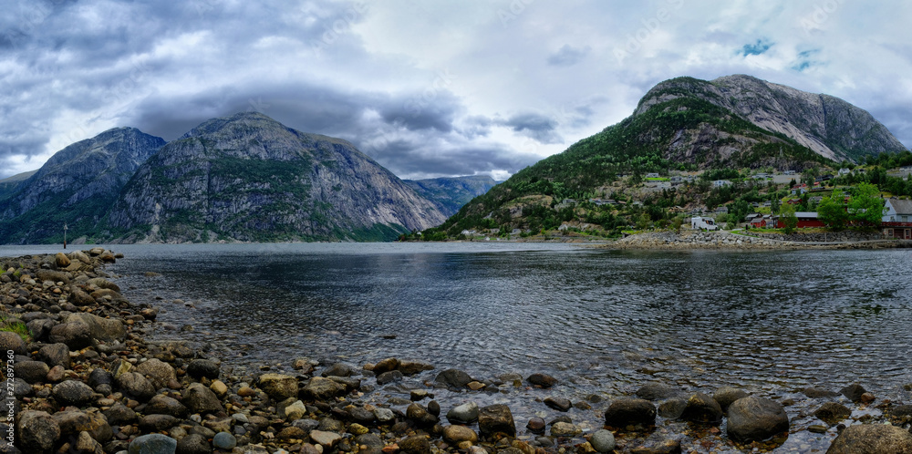 An endless chain of mountains reflecting itself in a calm water of Eidfjord. Taller parts of the mountains are partially covered with snow. Cloudy weather. Romantic landscape
