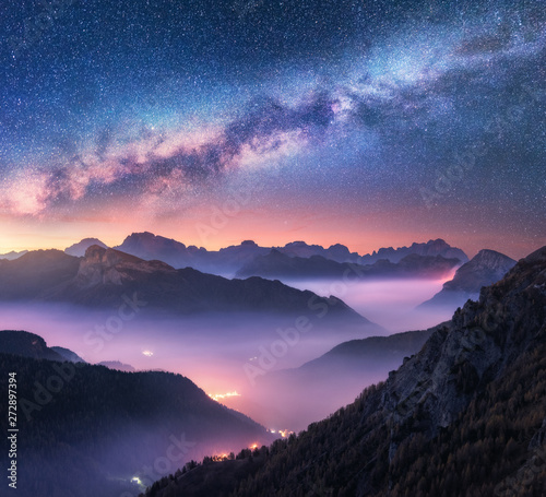Milky Way over mountains in fog at night in summer. Landscape with alpine mountain valley, purple low clouds, colorful starry sky with milky way, city illumination. Passo Giau, Dolomites, Italy. Space