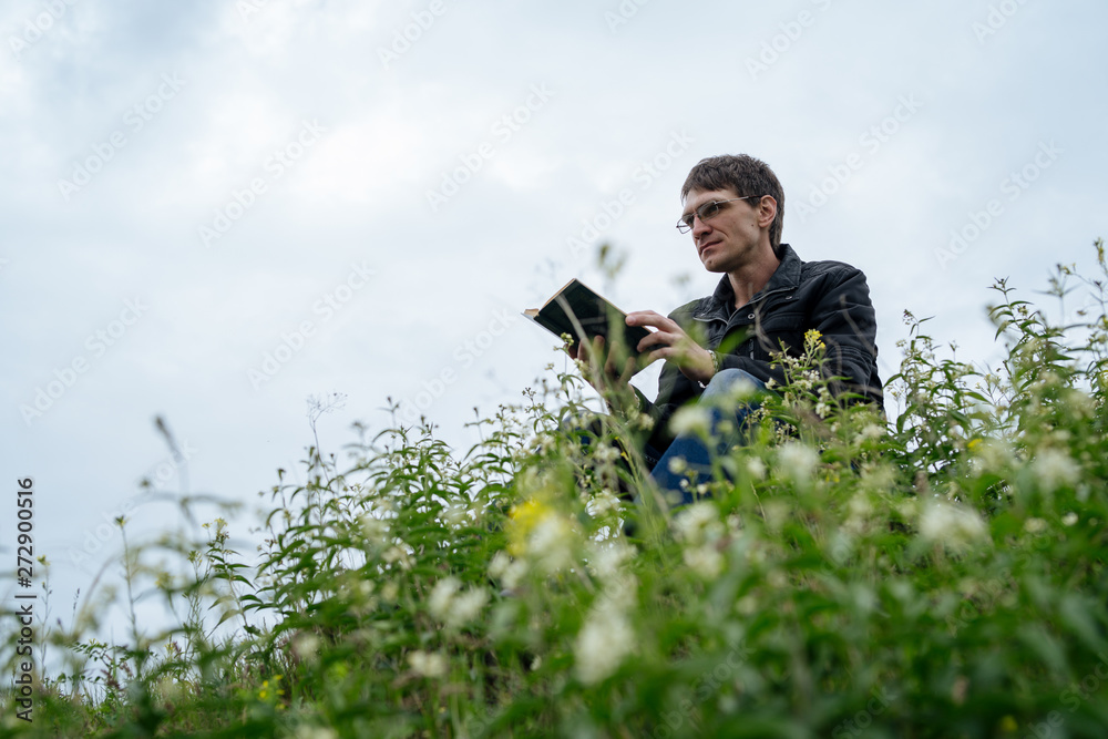 Man reading book in green hilly field. Side view of young handsome man in glasses and warm jacket reading book in open space on background of green hills Man reading book in green hilly field. Side view of young handsome man in glasses and warm jacket reading book in open space on background of green hills
