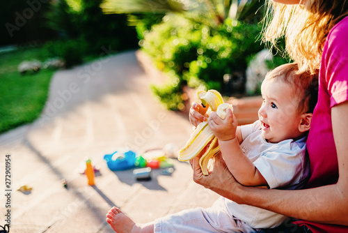 Adorable baby with a dirty face nibbling a banana sitting with his mother in his yard.