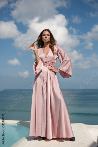 Beautiful young woman model in a long silk pink dress stands against the blue sky and sea view. fashion posing. white clouds in the background
