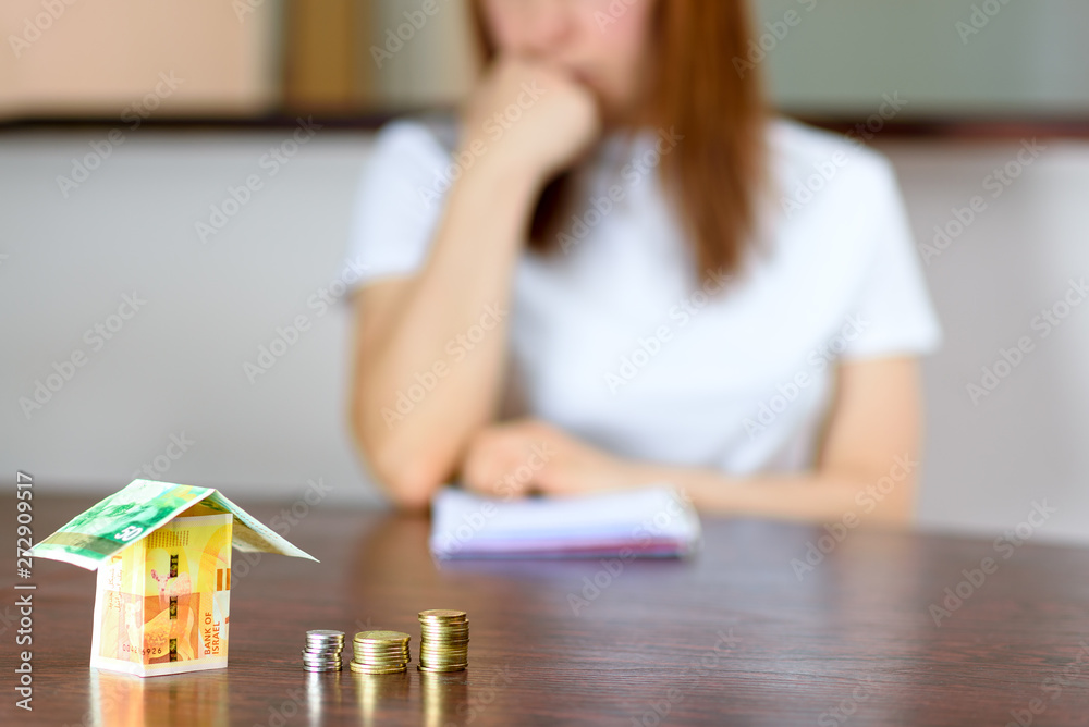 Middle age Woman With Model House from Israel Currencies new israeli shekel On wooden Table. Woman relaxing at home, building house from Israel 100 and 50 banknotes.Selective focus on money.
