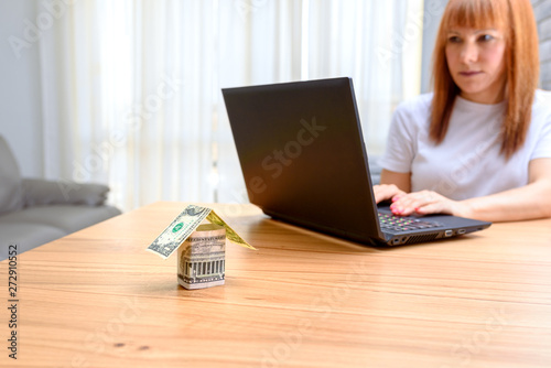 Money house from dollar banknote on wooden table.Middle age worried woman using laptop sitting in the living room at home.Concept of buying a home, real estate activity, looking new homes for sale.