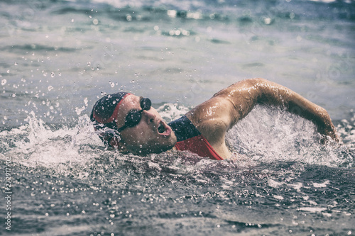 Triathlon race swim swimmer swimming freestyle crawl in ocean. Professional male triathlon swimmer wearing cap  goggles and red triathlon tri suit training for ironman breathing out of water.